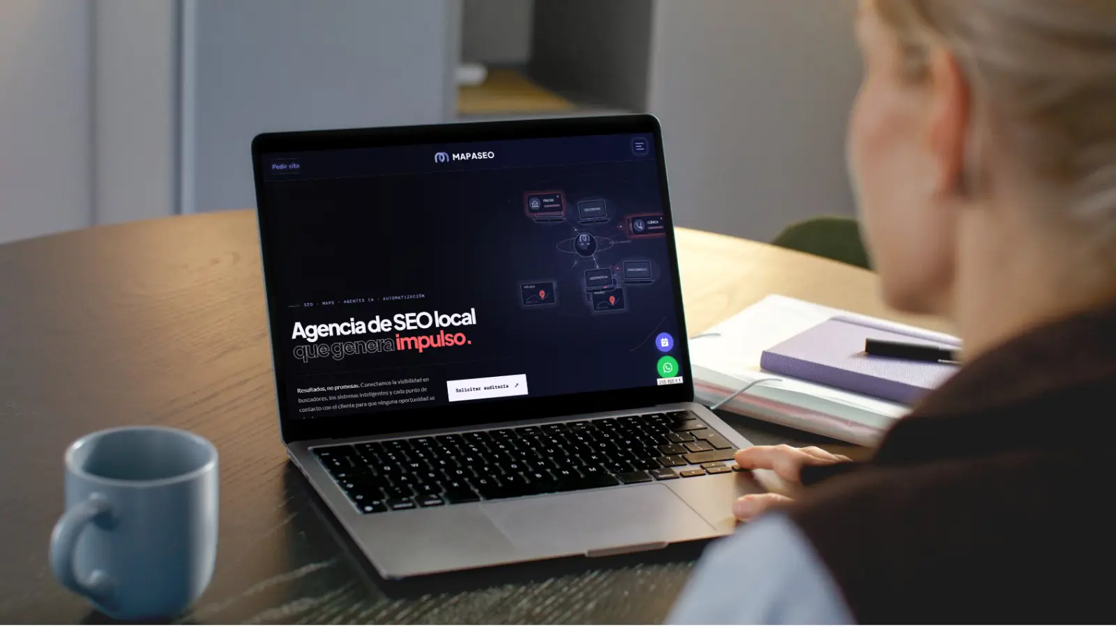 Una persona trabaja en un portátil con una web de agencia de SEO local abierta en pantalla, sobre una mesa de madera con una taza y un cuaderno.